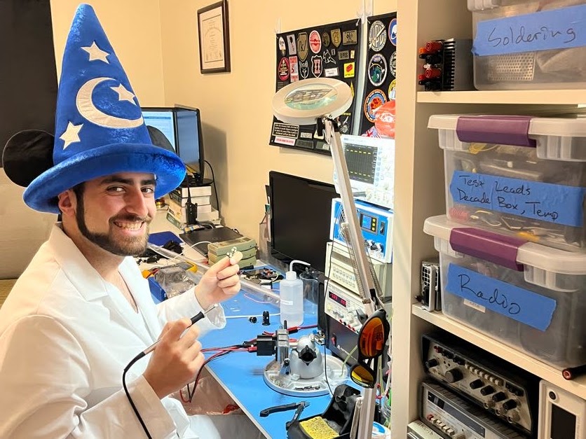Me at my electronics bench wearing a ridiculous lab coat and Mickey Mouse sorcerer hat outfit holding a soldering iron and smiling like an idiot